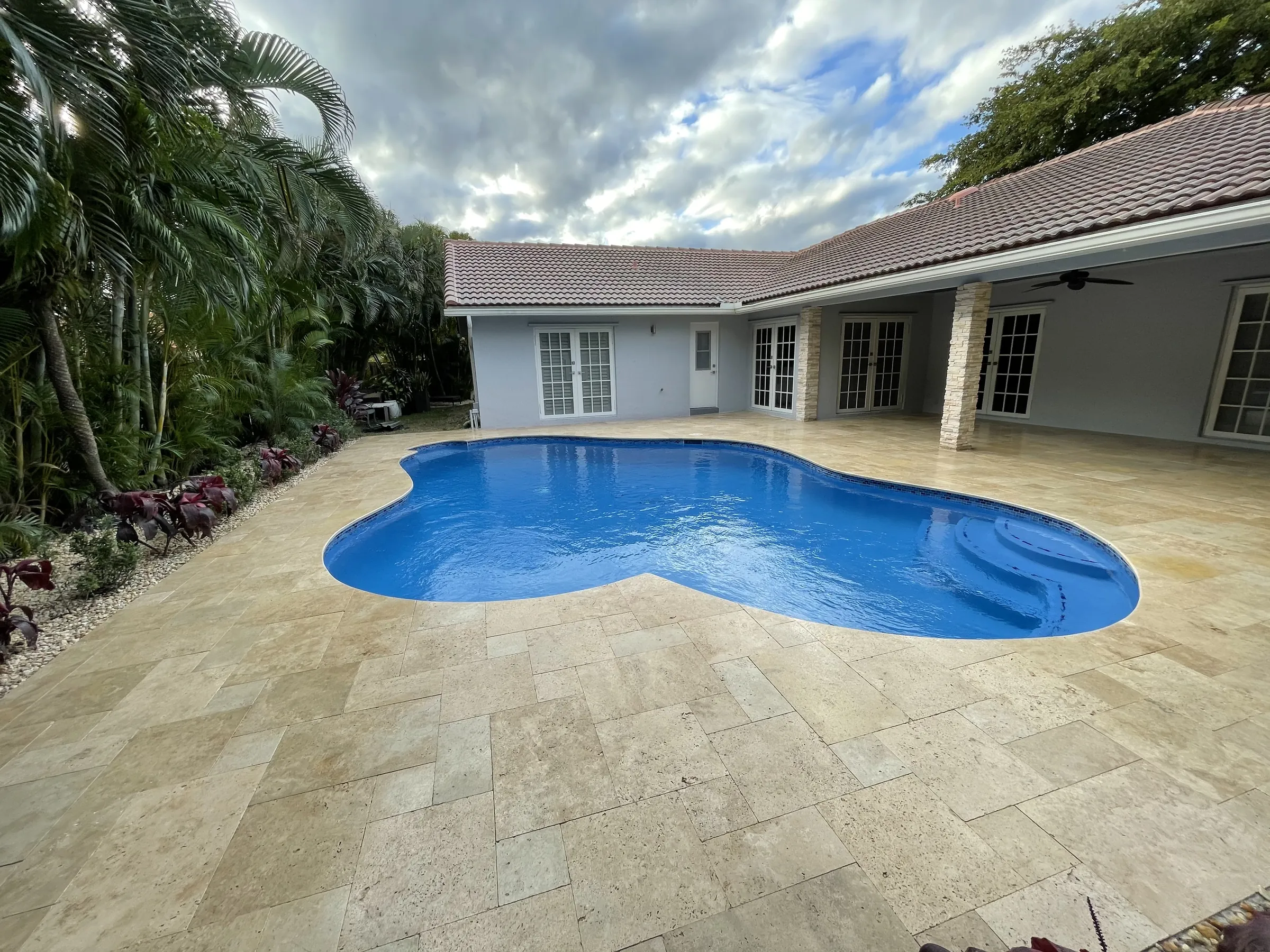 Renovated freeform pool with a deep blue finish and a travertine-style paver deck.